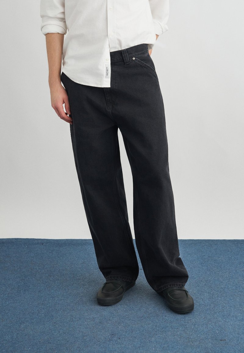 Uomo con pantaloni neri larghi, scarpe nere e camicia bianca a maniche lunghe con maniche arrotolate, in piedi su un tappeto blu.