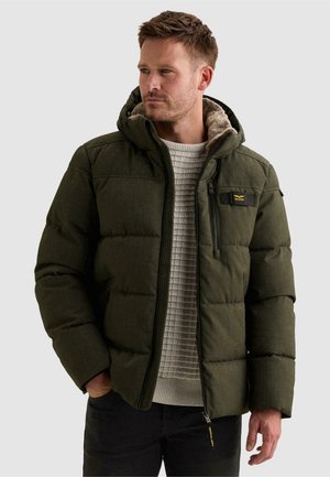Homme portant une veste matelassée vert olive avec une capuche doublée de fourrure par-dessus un pull en maille gris clair, regardant légèrement sur le côté.