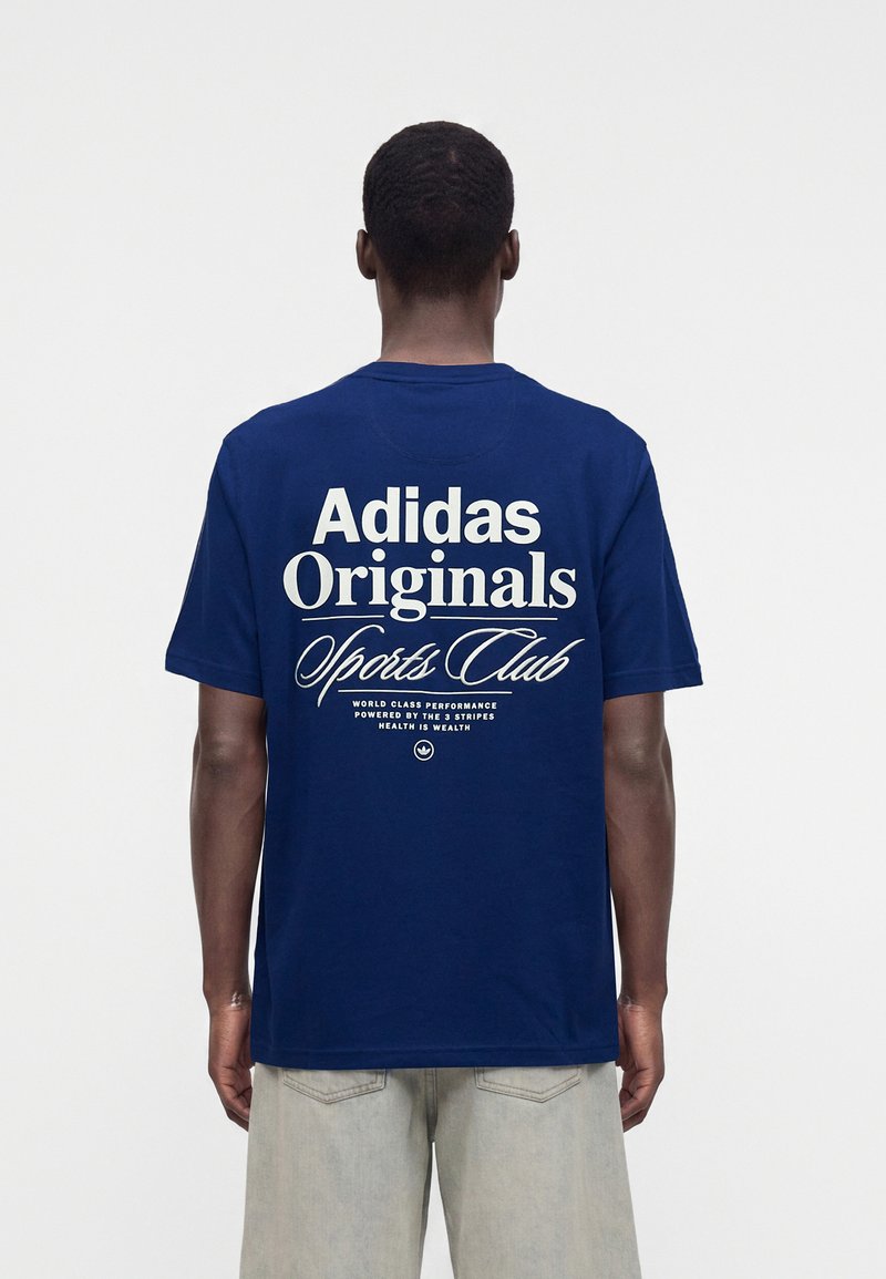 Άνδρας που φοράει μπλε t-shirt Adidas Originals Sports Club με λευκό κείμενο στην πλάτη, συνδυασμένο με ανοιχτόχρωμο παντελόνι.