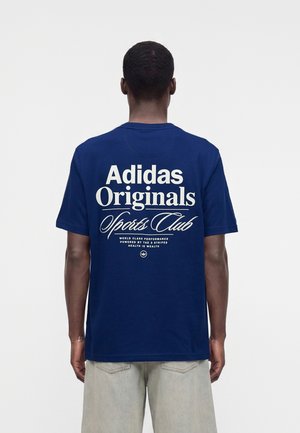 Άνδρας που φοράει μπλε t-shirt Adidas Originals Sports Club με λευκό κείμενο στην πλάτη, συνδυασμένο με ανοιχτόχρωμο παντελόνι.