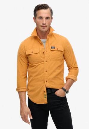 Long-sleeve, button-up shirt in mustard yellow cotton with two chest pockets and a label. Worn with black pants and a watch, collar raised.