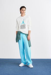 Balta garām piedurknēm krekls ar tirkīza logotipu un melnām svītrām; apvienots ar gaiši ziliem džogeriem; kājās baltas sporta kedas.