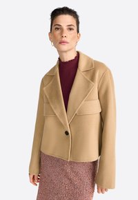 Kort beige wolblend jack met een uitslagkraag, enkele zwarte knop en twee voorzakken. Model draagt een bordeauxrood top en een gestreepte rok.