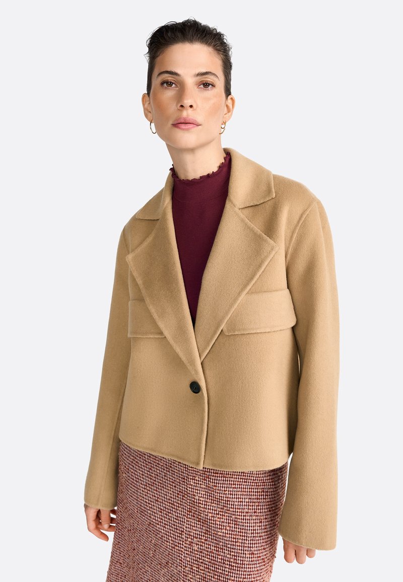 Kort beige wolblend jack met een uitslagkraag, enkele zwarte knop en twee voorzakken. Model draagt een bordeauxrood top en een gestreepte rok.