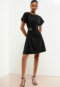 Schwarzes Kleid mit kurzen Ärmeln, einer taillierten Taille mit einem goldenen ovalen Gürtel und einem fließenden Rock. Glatte Stoffoberfläche und elegante Silhouette.