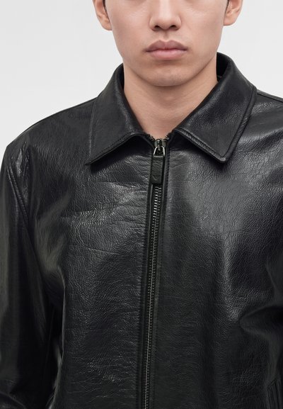 Man wearing a black zip-up leather jacket with a pointed collar, shown from shoulders to chin against a plain light background.