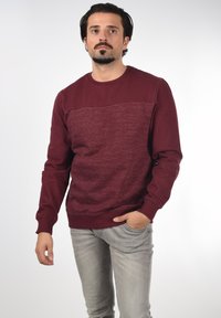 Bordeauxfarbener Pullover mit einem strukturierten Strickmuster im oberen Bereich, gerippten Bündchen und klassischem Rundhalsausschnitt. Getragen mit hellgrauen Jeans.