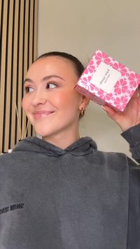 Różowe pudełko na prezent w kwiatowy wzór z białą etykietą, trzymane na tle szarej bluzy z kapturem. Pudełko zdobią żywe kwiaty i geometryczny wzór.