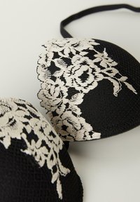 Svart bh i texturerat tyg, med delikat vit blommig broderi på kuporna och justerbara svarta banden.