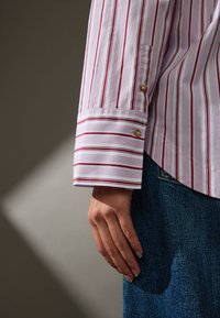 Camicia bianca a righe rosse, con polsini abbottonati dotati di bottoni dorati, indossata sopra denim blu. Tessuto di cotone liscio, vestibilità sartoriale.