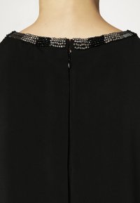 Svart plagg med en slät textur, med en pärlbeströdd halsringning i silver- och svartnyanser, samt en dragkedjeslutning bak.