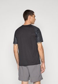 Sort atletisk shirt med teksturerede sorte mønstrede ærmer, rund halsudskæring og en løs pasform. Bæres med grå shorts.