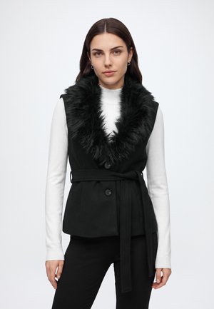 Femme portant une veste noire sans manches ceinturée avec un large col en fausse fourrure sur un haut blanc à manches longues, sur un fond uni.