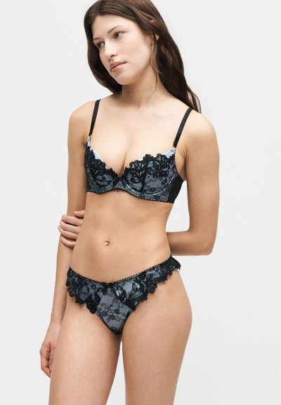 Νέα γυναίκα που φοράει σετ από μαύρο δαντελένιο σουτιέν και εσώρουχο, στέκεται με το ένα χέρι πίσω από την πλάτη της, κοιτάζοντας ελαφρώς προς τα πλάγια.