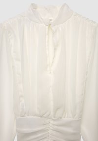 Camicia bianca con colletto alto, caratterizzata da pieghe verticali, apertura a goccia sulla scollatura e dettagli arricciati lungo il corpetto.