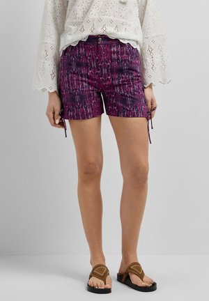 Femme portant un short violet à motifs avec des liens latéraux, un chemisier blanc en broderie anglaise et des sandales marron, debout devant un fond gris uni.