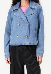 Chaqueta de mezclilla azul clara con un diseño de doble botonadura, solapas anchas, botones plateados y dos bolsillos laterales. Combinada con pantalones de rayas negras.