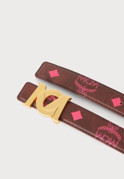 Ceinture en cuir marron avec motifs de diamants roses et logos MCM, avec une grande boucle à logo MCM dorée sur un fond blanc uni.
