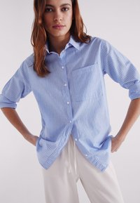 Light blue striped button-up shirt with a collar, relaxed fit, and a chest pocket, paired with light-colored pants. Smooth fabric texture.