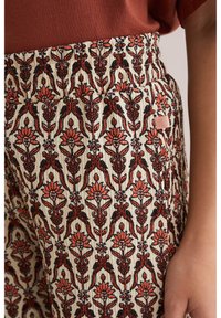 Krēmkrāsas kokvilnas bikses ar ziedu rakstu, kurās ir sarkani un melni dizaini, ar elastīgu jostasviedi un sānu kabatu. Tekstūrēts, ikdienišķs stils.