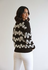 Kobieta o ciemnych włosach do ramion, ubrana w czarny sweter z białymi, falistymi paskami i białe spodnie, spoglądająca przez ramię.