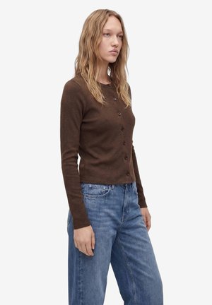 Ung kvinde med langt, bølget blondt hår iført en brun knappet cardigan med lange ærmer og løse blå jeans, stående sidelæns.