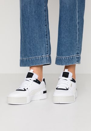 Baskets blanches Puma avec des accents noirs portées avec un jean bleu court, debout sur un sol gris clair devant un fond uni.