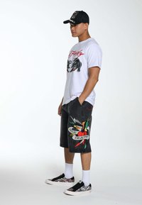 Shorts di jeans neri con grafiche ispirate ai tatuaggi colorati, abbinati a una t-shirt bianca con grafica e scarpe da ginnastica con fantasia. Un cappellino nero completa l'outfit.