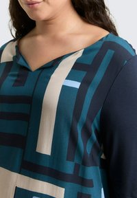 Multikleurige dames top met een V-hals, geometrische patronen in teal, marineblauw en beige, met een zachte stofstructuur en een gestructureerde pasvorm.