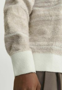 Beige textuurtrui met een zachte, pluizige afwerking. Voorzien van witte geribbelde manchetten, die een subtiel geweven patroon tonen.