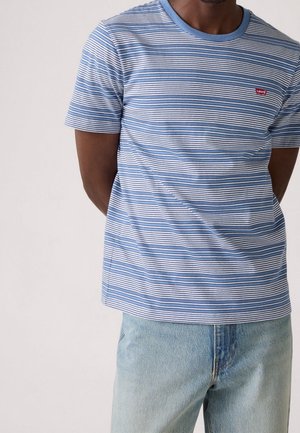 Mand iført en blå og hvid stribet Levi's t-shirt, let stukket ned i lyseblå jeans, hænderne bag ryggen, stående mod en ensfarvet baggrund.