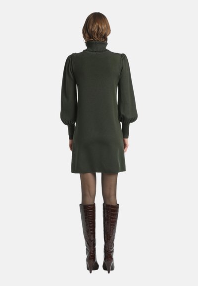 Grünes Strickkleid mit hohem Kragen, Ballonärmeln und gerader Silhouette, kombiniert mit hochglänzenden burgunderfarbenen, krokodil-prägierten Stiefeln.