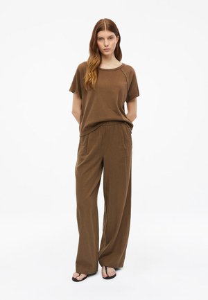 Femme aux longs cheveux bruns portant une chemise marron ample à manches courtes et un pantalon à jambes larges, debout les mains dans le dos.
