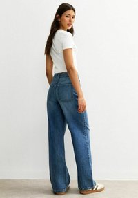 Mujer con cabello oscuro y largo que lleva una blusa blanca de manga corta, unos pantalones vaqueros anchos de tiro alto en azul y zapatillas blancas con rayas marrones.
