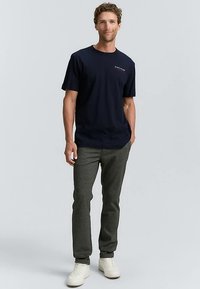 Temno modra bombažna majica s kratkimi rokavi, kombinirana s sivimi ohlapenimi hlačami in belimi supergami, prikazana na modelu na svetlem ozadju.