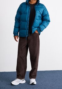 Tealblå vadderad jacka med dragkedja och ribbade muddar, matchad med bruna manchesterbyxor och vita sneakers. Texturerad och lager-på-lager design.