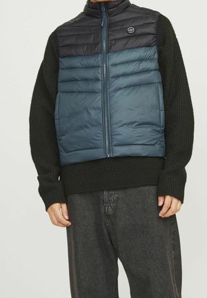 Uomo che indossa un maglione nero lavorato a maglia e un gilet imbottito smanicato blu e nero con zip, abbinato a pantaloni grigio scuro, in piedi davanti a uno sfondo uniforme.