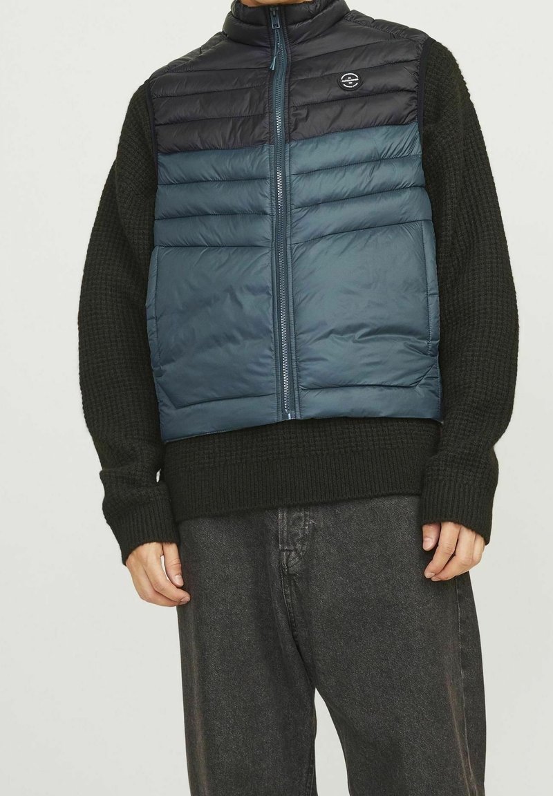 Man wearing black knitted sweater and zip-up sleeveless blue and black padded vest, paired with dark gray pants, standing against plain background.