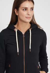 Svart hettegenser med glidelås, foret hette, kremfargede snor, og brune lærdetaljer ved hals og lommer; myk stofftekstur.