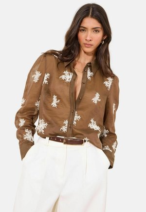 Vrouw met lang donker haar, gekleed in een bruin blouse met knopen en witte bloemrijke borduursels, en een witte broek met hoge taille en een bruine riem.