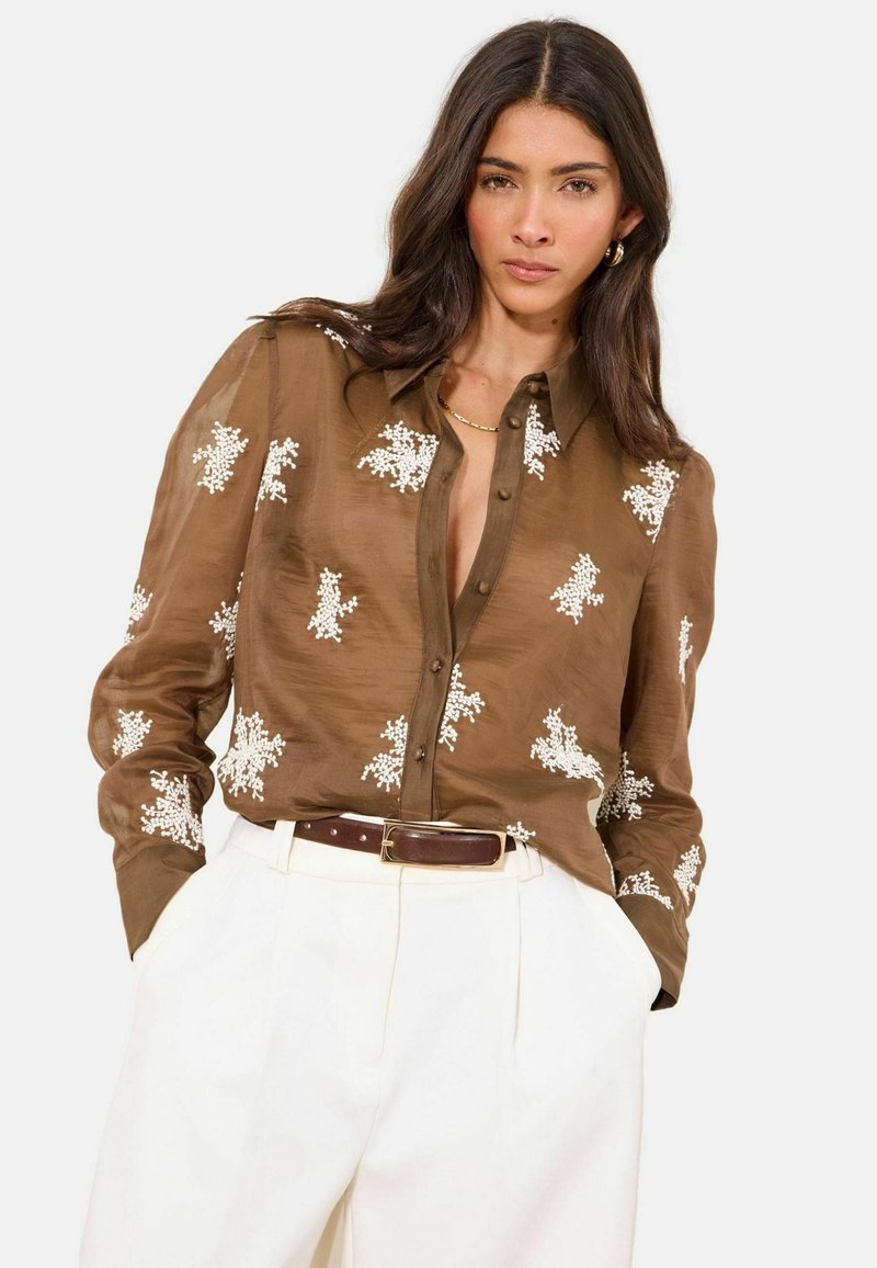 Žena s dlouhými tmavými vlasy, oblečená v hnědé košili na knoflíky s bílou květinovou výšivkou a vysokým pasem bílých kalhot s hnědým opaskem.