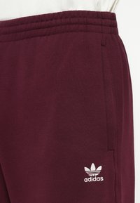 Tuvumā redzami bordo Adidas sporta bikses ar elastīgu jostu un baltu Adidas trifoliju logotipu blakus sānu kabatai.