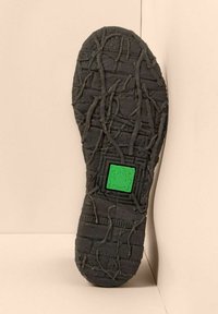Suela de zapato negra con patrones de pisada irregulares y un logo cuadrado verde con una rana en el centro sobre un fondo beige.