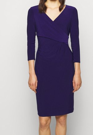 Mujer con un vestido envolvente ajustado de manga larga, largo hasta la rodilla, de color púrpura oscuro, con escote en V, sobre un fondo claro y liso.