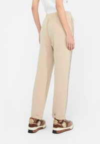 Pantalons de jogging beiges avec une taille élastique, présentant une bande latérale brillante. Portés avec des baskets brunes, affichant un talon rembourré et un dessus texturé.