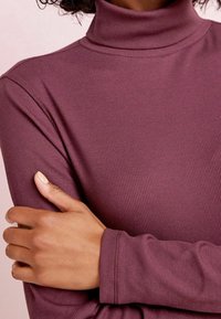 Haut à col montant à manches longues en tissu côtelé mauve profond. Présente un ajustement cintré avec un col haut et une texture douce.