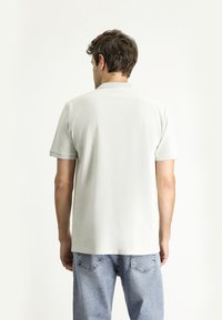 Polo grigio chiaro con colletto e maniche corte, caratterizzato da una texture liscia. Abbinato a jeans in denim blu chiaro.