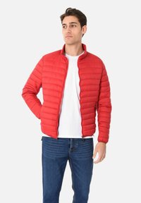 Giacca a vento rossa con texture trapuntata e colletto alto, dotata di zip frontale e tasche laterali. Indossata sopra una camicia bianca e jeans blu.