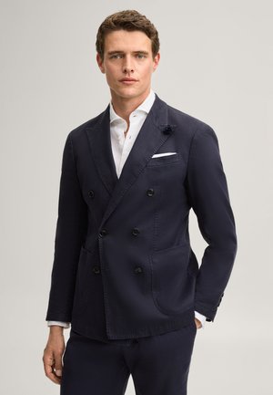 Mann trägt marineblauen zweireihigen Blazer über weißem Hemd, steht vor einfarbig hellem Hintergrund, schaut geradeaus mit neutralem Gesichtsausdruck.