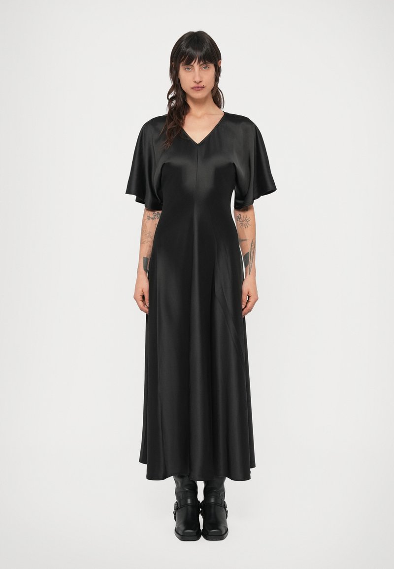 Čierne saténové šaty s výstrihom do V, krátkymi nadýchanými rukávmi a splývavým áčkovým strihom, nosené s čiernymi členkovými čižmami. Hladká textúra zdôrazňuje eleganciu.